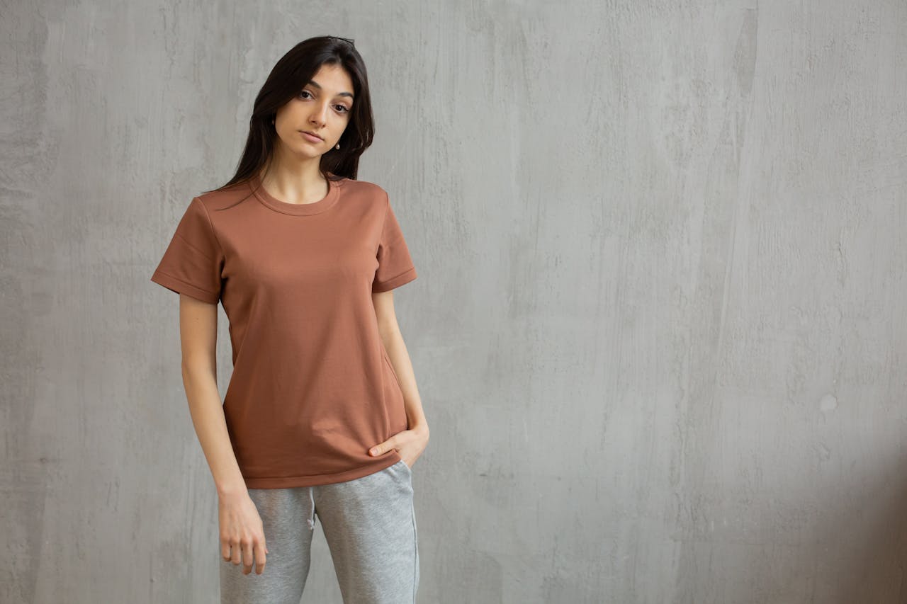 Serious female with long hair wearing t shirt and sweatpants standing against gray wall and looking at camera